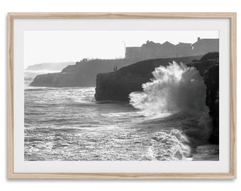 May include: Black and white framed print of a coastal scene. A large wave crashes against a cliff, creating a dramatic splash. Buildings are visible on the cliff's edge, and the ocean stretches to the horizon.