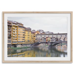 Op de afbeelding: Een foto van de Ponte Vecchio-brug in Florence, Italië. De brug is bezaaid met winkels en gebouwen, en de rivier de Arno stroomt eronderdoor. De gebouwen zijn een mix van geel, oranje en rood.