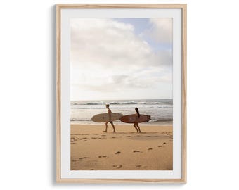 May include: A framed print featuring two people walking on a sandy beach, each carrying a surfboard. The surfboards are different colors, one light brown and the other reddish-brown. The ocean and sky are visible in the background.