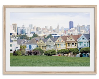 Könnte beinhalten: Eine Reihe von bunten viktorianischen Häusern mit Blick auf die Skyline von San Francisco im Hintergrund. Die Häuser sind in Blautönen, Grün, Gelb und Rosa gestrichen. Die Skyline umfasst die ikonische Transamerica-Pyramide.
