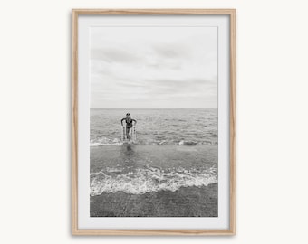May include: Black and white framed photograph of a person in the ocean. The person is standing in the water, holding onto a set of parallel bars. The sky is overcast, and the water is choppy.