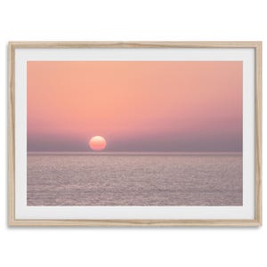 Puede incluir: Una impresión enmarcada que muestra una serena puesta de sol sobre el océano. El cielo transiciona de un rosa suave a un púrpura más profundo, con el sol apareciendo como un orbe brillante. El agua está tranquila, reflejando los colores del cielo. El marco es de madera clara.
