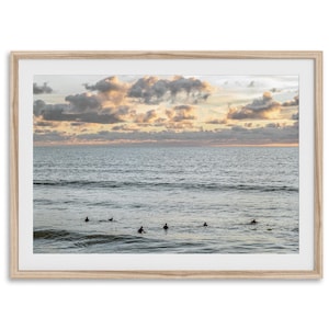 May include: A framed print of a seascape featuring surfers in the ocean at sunset. The sky is filled with peach and gray clouds, and the water reflects the colors of the sky. The frame is a light wood color.