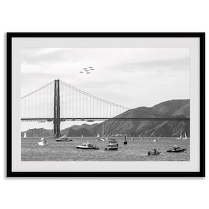 Może przedstawiać: Czarno-biała oprawiona grafika przedstawiająca most Golden Gate w San Francisco. Obraz przedstawia most nad wodą z łodziami i samolotami na niebie. Grafika jest w czarnej ramie.