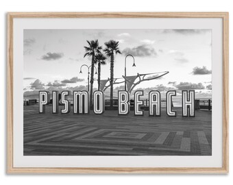 Puede incluir: Impresión enmarcada en blanco y negro con las palabras "PISMO BEACH" en letras grandes e iluminadas. Se ven palmeras y un muelle al fondo, con un cielo nublado arriba. La imagen es una vista panorámica de una playa.