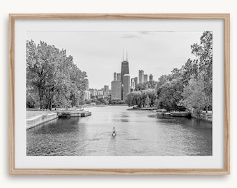 May include: Black and white framed print of a river scene with a person rowing a boat. The Chicago skyline is visible in the background, framed by trees on either side of the river. The print is in a light-colored wooden frame.