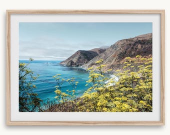May include: A framed landscape print featuring a coastal scene. The image showcases a vibrant blue ocean, rugged mountains, and yellow wildflowers. The sky is a soft blue, and the overall aesthetic is serene.