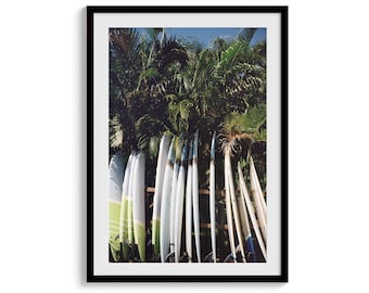 May include: A framed vertical wall art print featuring a row of multiple white and light green surfboards standing upright against a wooden rack, positioned in front of dense, dark green palm tree foliage under a clear blue sky.