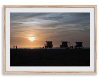 May include: A framed landscape photograph featuring three lifeguard towers silhouetted against a sunset sky over a beach. The scene includes dark figures of people standing on the sand near the ocean horizon under a cloudy, golden-hued sky.