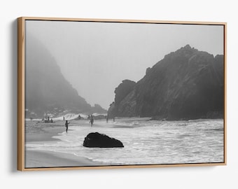 May include: A black and white photograph of a beach with rocky cliffs in the background. The beach is empty except for a few people walking along the shore. The water is calm and the sky is overcast.
