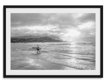 Puede incluir: Impresión enmarcada en blanco y negro de un surfista saliendo del océano con una tabla de surf. La playa y el océano están en primer plano, con montañas y un cielo nublado en el fondo. La imagen está en escala de grises.