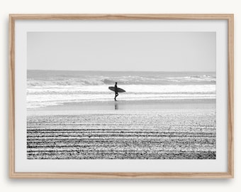May include: Black and white framed print of a surfer walking on a beach, carrying a surfboard. The ocean waves are visible in the background, and the sand is textured with lines. The image is a grayscale photograph.
