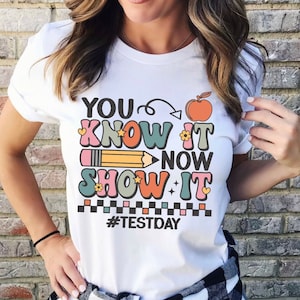 May include: White t-shirt with the text "YOU KNOW IT NOW SHOW IT" in colorful retro font, a pencil, and an apple graphic. The shirt also has the hashtag #TESTDAY.