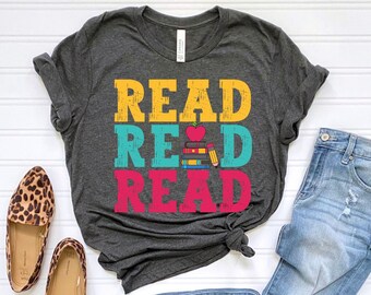 May include: A dark grey t-shirt with the words "READ READ READ" in yellow, blue, and pink. The word "READ" in the middle has a stack of books and a red apple with a heart on it.