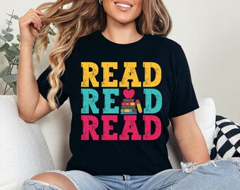 May include: A black t-shirt with the words "READ READ READ" in colorful, distressed lettering. A stack of books with an apple on top and a pencil are in the middle of the words.