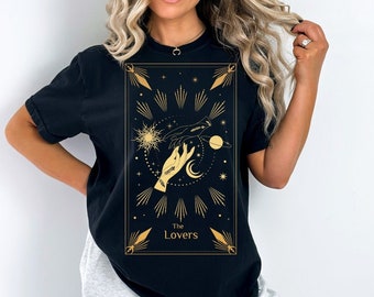 May include: Black t-shirt with a gold graphic of the Lovers tarot card. The card features two hands reaching towards each other, a sun, a moon, and stars. The text "The Lovers" is printed below the graphic.
