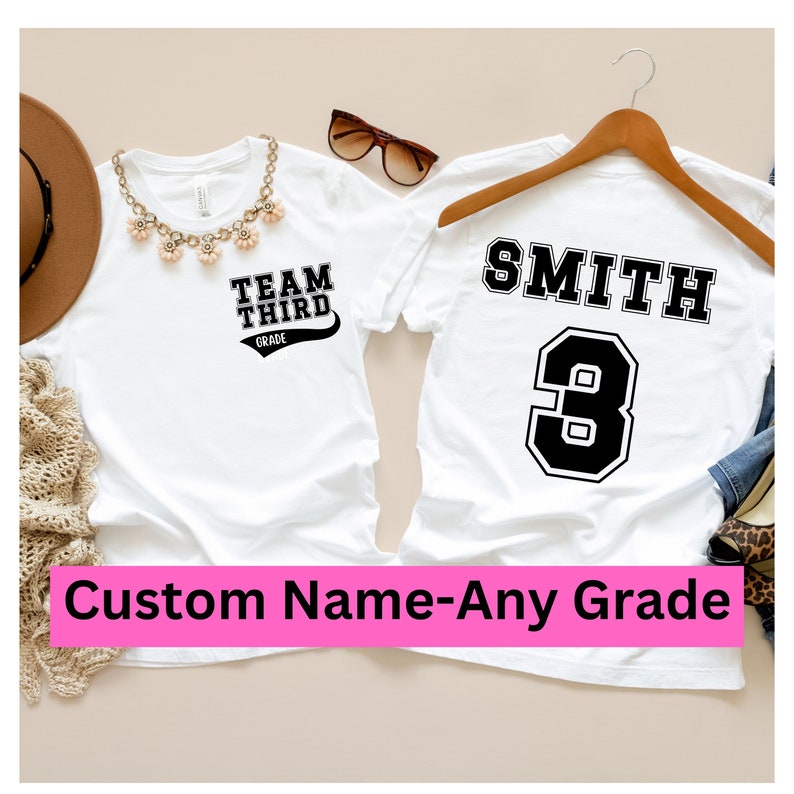 K&ouml;nnte beinhalten: Zwei wei&szlig;e T-Shirts, eines mit dem Text "TEAM THIRD GRADE" und das andere mit dem Text "SMITH 3". Die Shirts liegen auf einem hellbraunen Hintergrund.