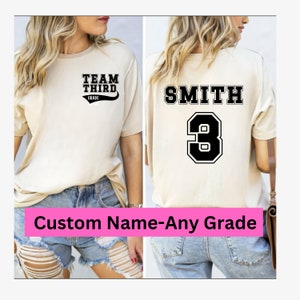 K&ouml;nnte beinhalten: Ein beiges T-Shirt mit schwarzem Text, der auf der Vorderseite "Team Third Grade" und auf der R&uuml;ckseite "Smith 3" lautet. Das Shirt wird von einer Person in Jeans-Shorts getragen.