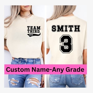 K&ouml;nnte beinhalten: Ein beiges T-Shirt mit schwarzem Text, der auf der Vorderseite "Team Third Grade" und auf der R&uuml;ckseite "Smith 3" lautet. Das Shirt wird von einer Person in blauer Jeans getragen.