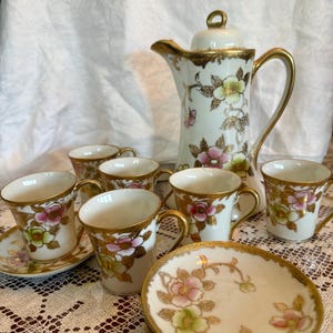 May include: A vintage porcelain tea set featuring a white teapot and matching cups and saucers. The set is decorated with pink, green, and gold floral patterns. The teapot has a gold handle and spout, and the cups have gold rims.