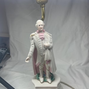 May include: A white porcelain lamp with a figurine of a man in a white and pink suit. The lamp has a gold base and a gold cord.