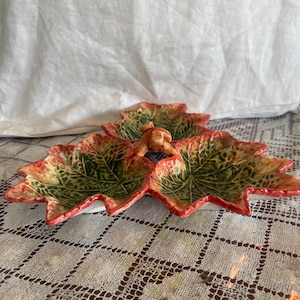 May include: A ceramic leaf-shaped serving dish with three sections. The dish has a green and yellow leaf design with red edges and a small handle. The dish is on a white and beige patterned tablecloth.