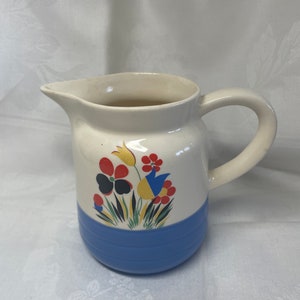 May include: A white ceramic pitcher with a blue band and a floral design featuring red, yellow, blue, black, and green flowers.