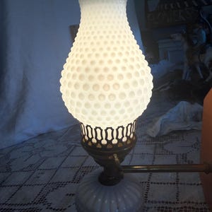 May include: A white hobnail glass lamp with a ruffled top and a decorative metal base. The lamp has a round, textured base and a long, brass arm. The lamp is lit, casting a warm glow.