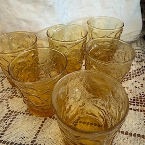 May include: Six amber-colored drinking glasses with a textured, organic pattern. The glasses are arranged on a white lace tablecloth. The glasses are made of glass and are likely vintage.