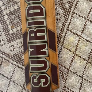 May include: A wooden cricket bat with the brand name "SUNRIDGES" in large, maroon letters. The bat has a light brown handle and a dark brown and yellow design. The bat is lying on a white lace tablecloth.