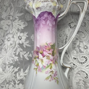 May include: A vintage porcelain teapot with a delicate floral design. The teapot features a white base with a purple and pink floral pattern. The handle is shaped like a scroll and has a gold trim. The lid has a scalloped edge and a gold knob.