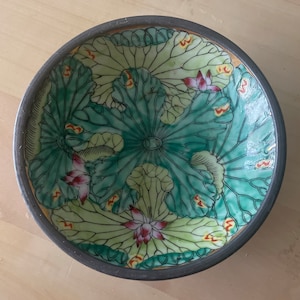 May include: A round ceramic bowl with a green and yellow lotus flower design. The bowl has a dark gray rim.