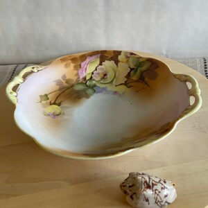 May include: A white porcelain bowl with two handles and a floral design featuring yellow and pink roses. The bowl is on a wooden surface.