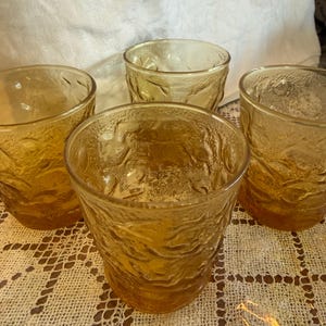 May include: Four amber-colored drinking glasses with a textured, wave-like pattern. The glasses are short and wide, with a slightly flared rim. They are arranged on a cream-colored, open-weave tablecloth.
