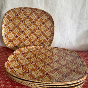 May include: A stack of oval-shaped plates with a repeating geometric pattern in shades of orange, yellow, and burgundy. The plates are stacked on a red patterned surface, with one plate slightly offset. The background is a white cloth.