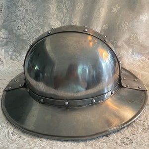 May include: A silver helmet with a wide brim and a rounded top. The helmet has a series of rivets around the brim and the top.