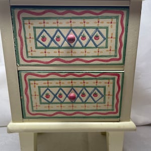 May include: A small white wooden chest with two drawers. The drawers have a colorful painted design with red, green, blue, and yellow accents. The chest has four short legs.