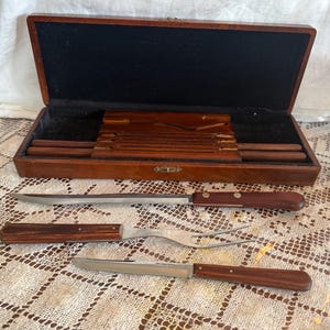 May include: A vintage wooden carving set with a wooden storage box. The set includes a carving knife, a carving fork, and a smaller knife. The handles are a dark brown color, and the blades are silver.