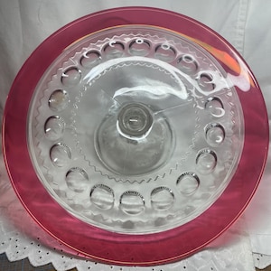 May include: A clear glass cake stand with a pink rim. The stand has a scalloped edge and a pattern of small circles around the rim.