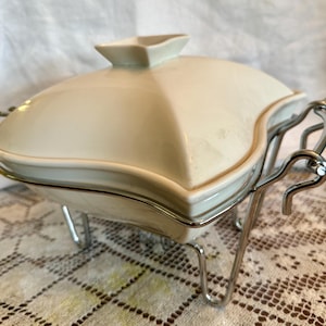 May include: Cream-colored, lidded serving dish with a square shape and a small, square handle on the lid. The dish sits in a chrome-colored metal stand with curved legs. The dish is likely made of ceramic.
