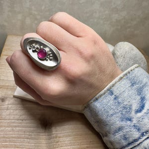 May include: A silver ring featuring a pink oval gemstone set within a textured oval bezel. The ring is worn on a hand with a denim jacket.