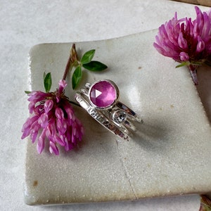 K&ouml;nnte beinhalten: Ein Set aus drei Silberringen mit einem rosafarbenen Edelstein in der Mitte. Die Ringe sind texturiert und haben eine geh&auml;mmerte Oberfl&auml;che. Der Edelstein ist rund und facettiert.