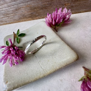 K&ouml;nnte beinhalten: Ein silberner Ring mit geh&auml;mmerter Textur. Der Ring liegt auf einer wei&szlig;en Keramikplatte mit rosa Bl&uuml;ten.