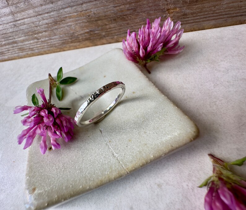 K&ouml;nnte beinhalten: Ein silberner Ring mit geh&auml;mmerter Textur. Der Ring liegt auf einer wei&szlig;en Keramikplatte mit rosa Bl&uuml;ten.