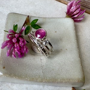 K&ouml;nnte beinhalten: Ein Satz aus drei silbernen Stapelringen mit einem rosafarbenen Edelstein und einem kleineren, klaren Edelstein. Die Ringe werden auf einer wei&szlig;en Keramikplatte pr&auml;sentiert.