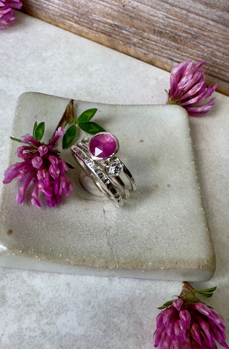 K&ouml;nnte beinhalten: Ein Satz aus drei silbernen Stapelringen mit einem rosafarbenen Edelstein und einem kleineren, klaren Edelstein. Die Ringe werden auf einer wei&szlig;en Keramikplatte pr&auml;sentiert.