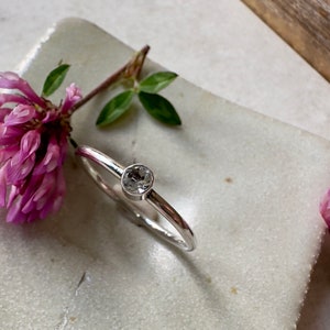 K&ouml;nnte beinhalten: Ein silberner Ring mit einem klaren, runden Edelstein in der Mitte. Der Ring liegt auf einer wei&szlig;en Oberfl&auml;che mit rosa Blumen auf beiden Seiten.