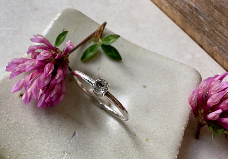 K&ouml;nnte beinhalten: Ein silberner Ring mit einem klaren, runden Edelstein in der Mitte. Der Ring liegt auf einer wei&szlig;en Oberfl&auml;che mit rosa Blumen auf beiden Seiten.