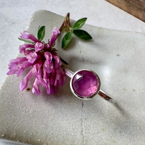 K&ouml;nnte beinhalten: Ein silberner Ring mit einem facettierten rosafarbenen Edelstein. Der Ring liegt auf einer wei&szlig;en Keramikplatte mit einer rosa Blume.