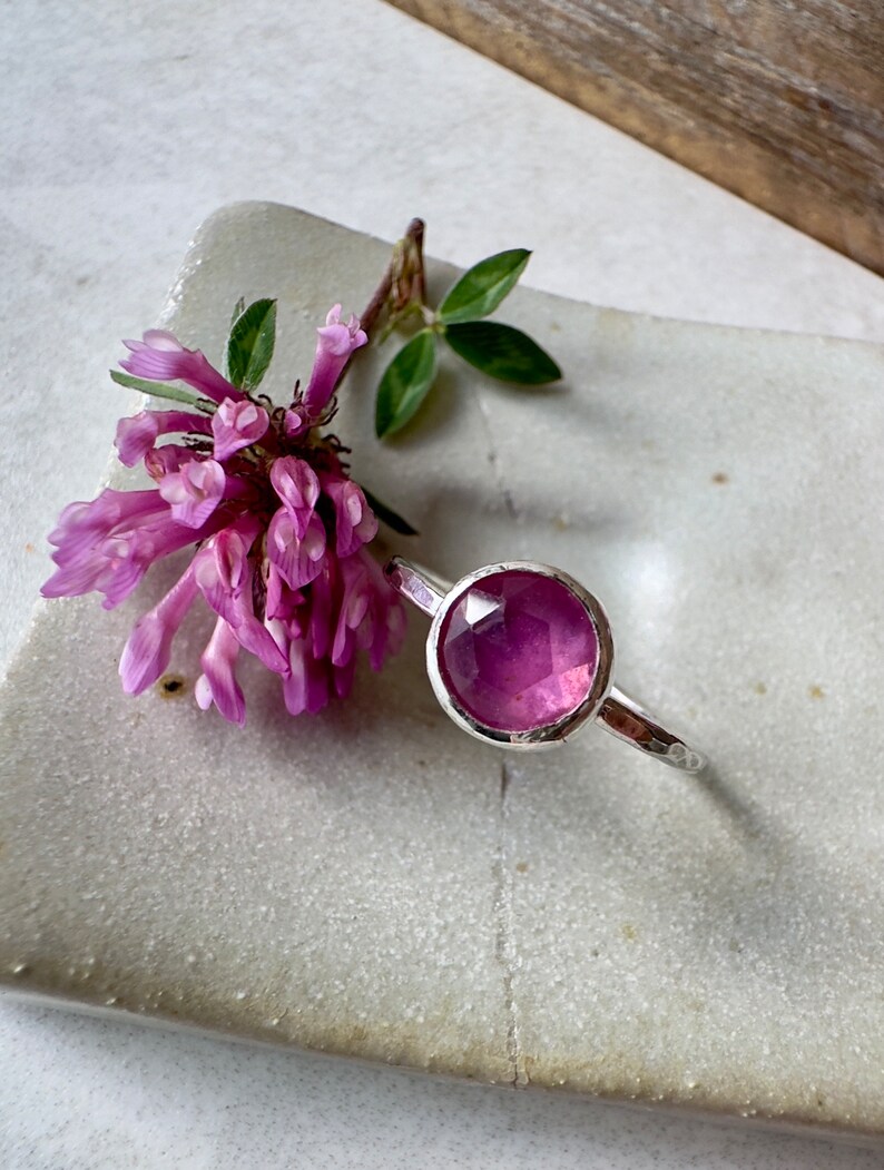 K&ouml;nnte beinhalten: Ein silberner Ring mit einem facettierten rosafarbenen Edelstein. Der Ring liegt auf einer wei&szlig;en Keramikplatte mit einer rosa Blume.